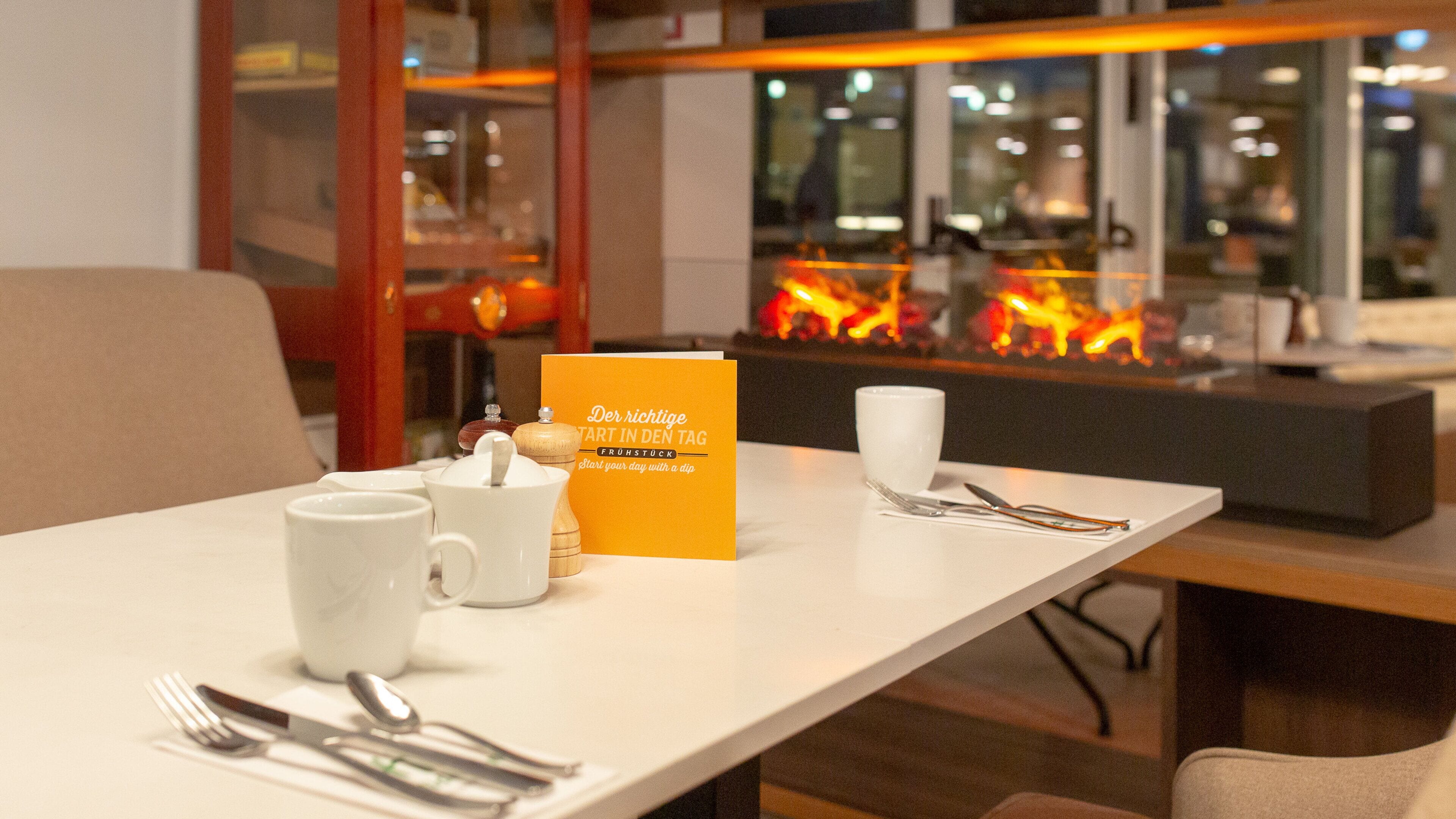 Een gedekte tafel met een koffiekop, theekop, suikerpot, lepel en servet. Er ligt een kaartje met tekst op en een haard in de achtergrond.