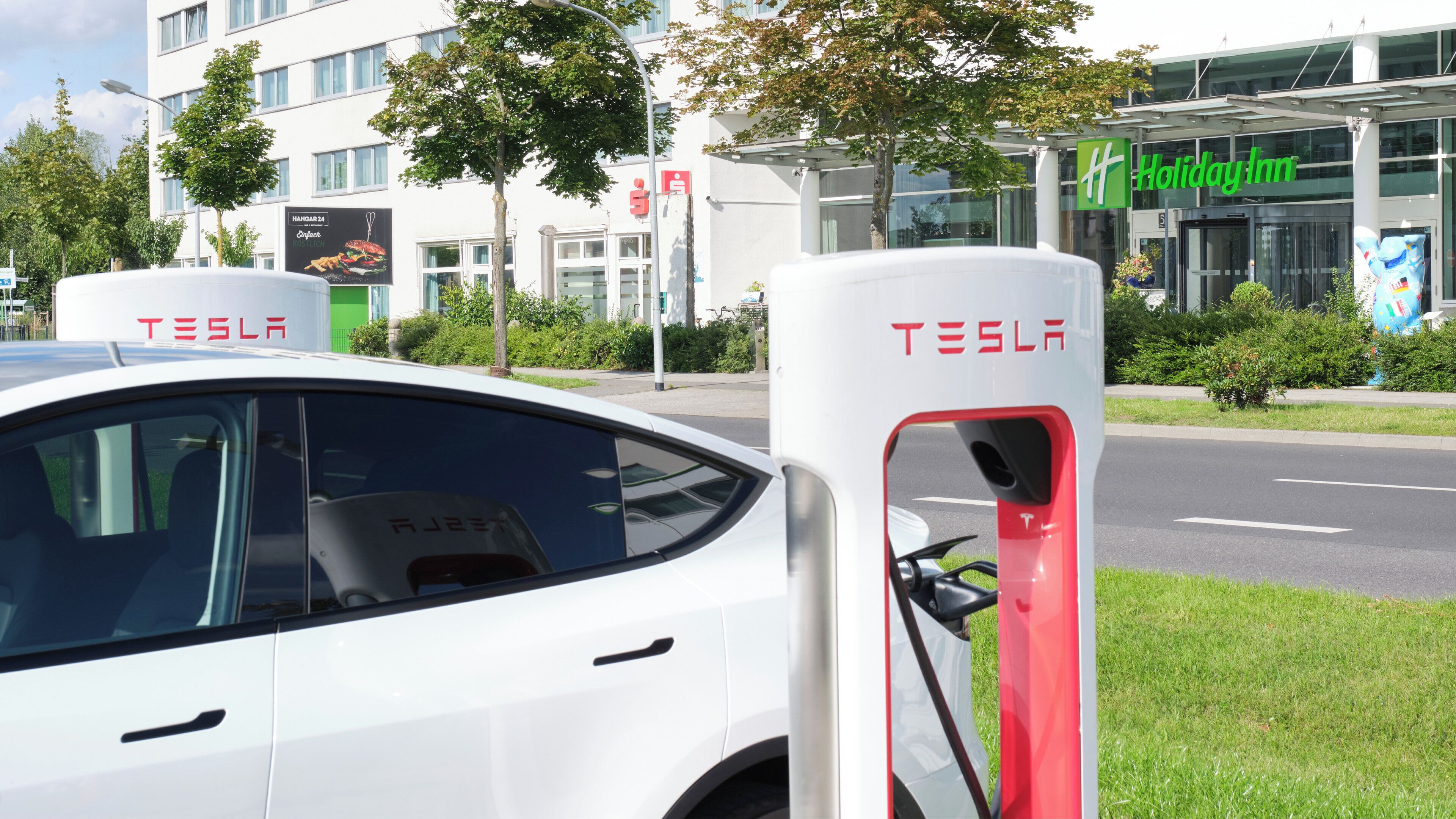 Un vehicle elèctric Tesla carregant-se en una estació davant d'un hotel Holiday Inn.