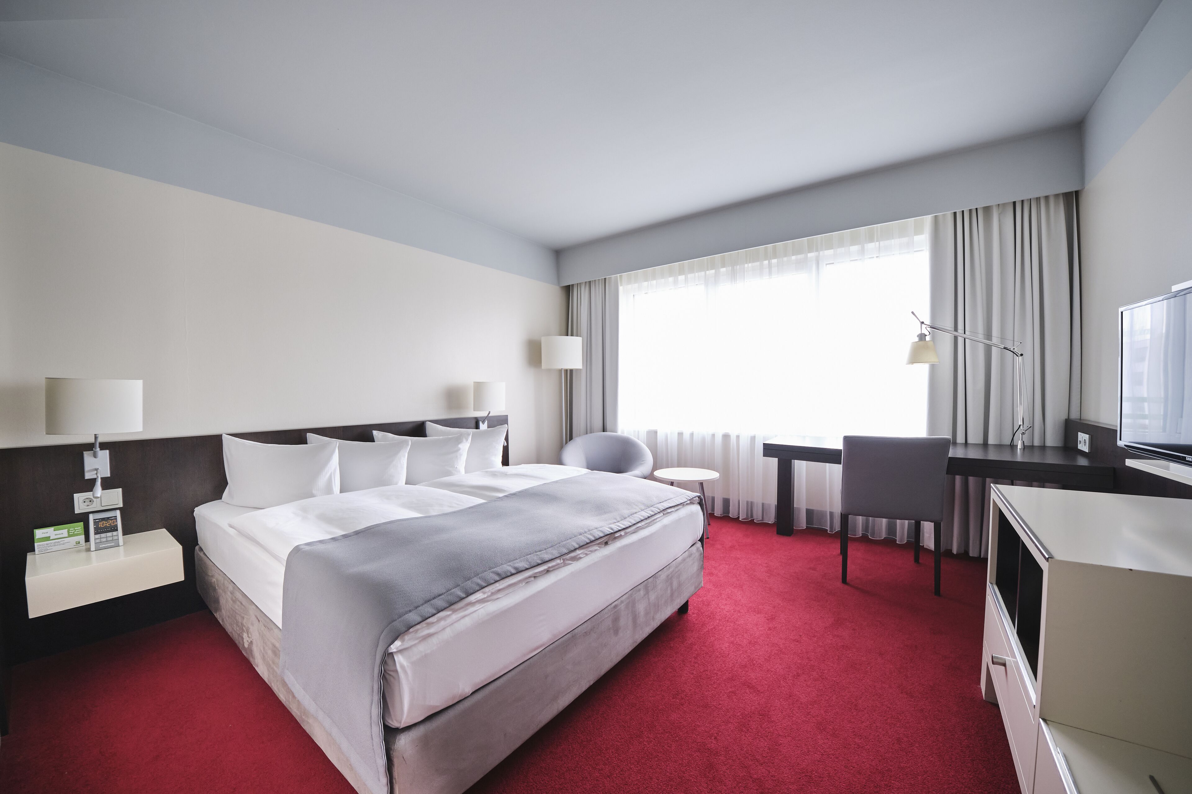 A hotel room with a large bed, a desk, a chair, a TV, and a window with curtains.