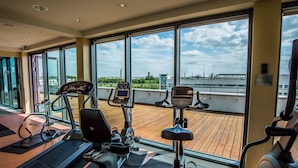 A gym with cardio machines and a wooden deck area, large windows with a view of the city. - Holiday Inn Berlin Airport - Conference Centre by IHG (Schoenefeld)