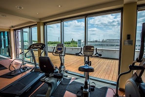 A gym with cardio machines and a wooden deck area, large windows with a view of the city. - Holiday Inn Berlin Airport - Conference Centre by IHG (Schoenefeld)