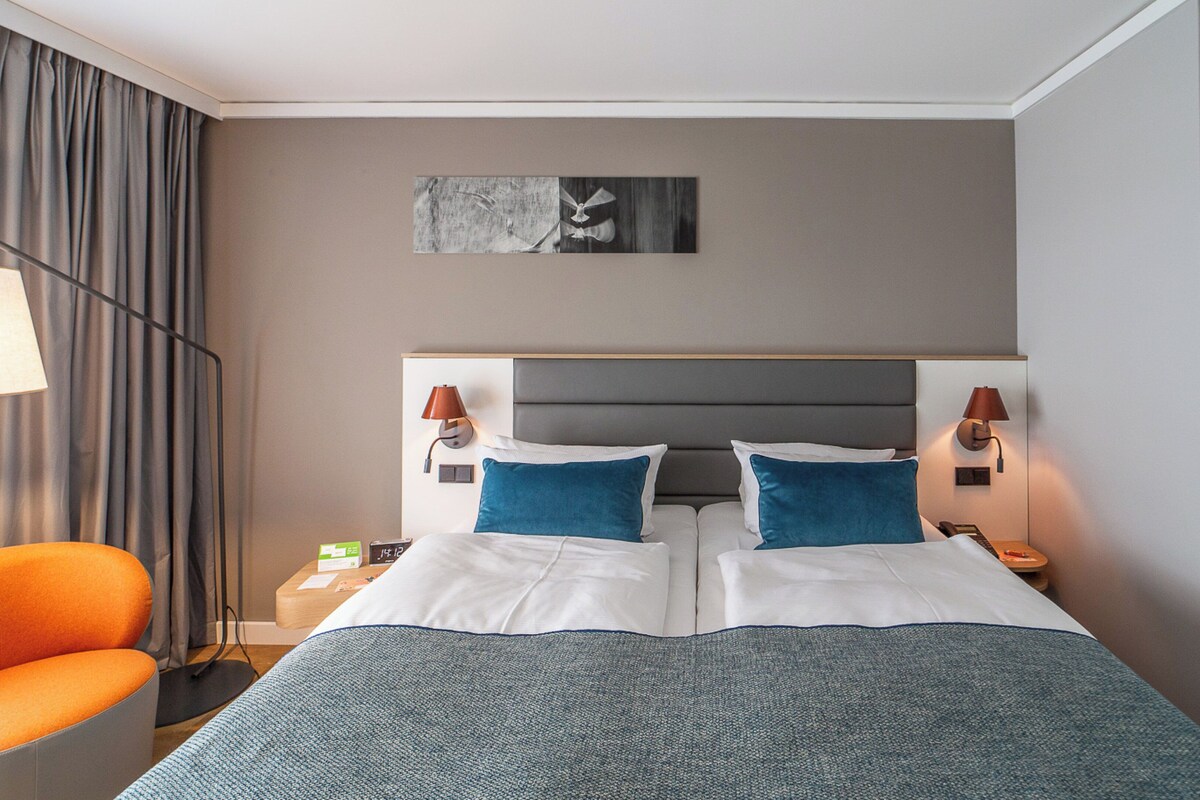 Una camera d'albergo con un letto grande, due comodini, una sedia, una lampada e un quadro appeso al muro.