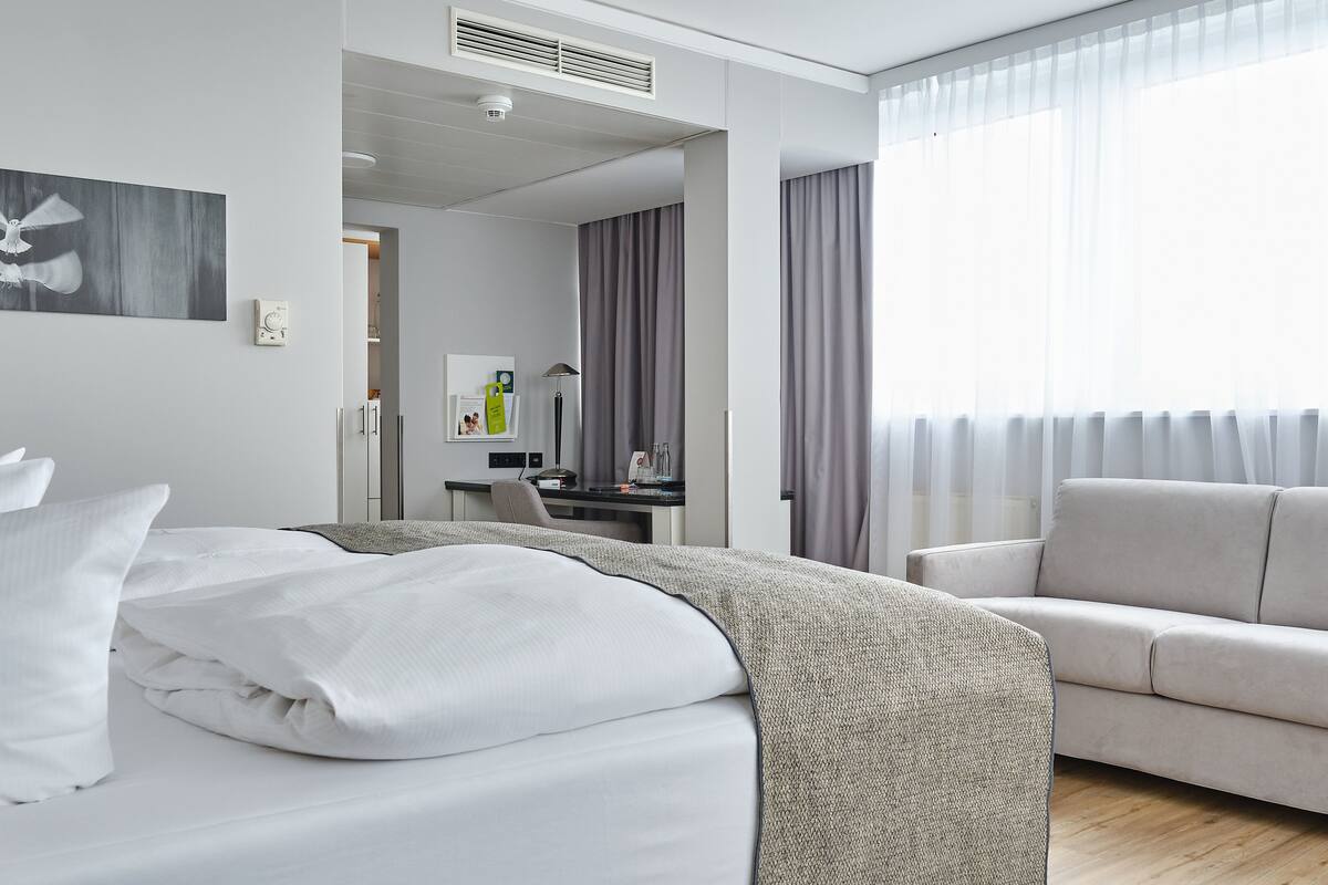 A modern hotel room with a neatly made bed, a sofa, a desk with a lamp, and a large window with curtains.