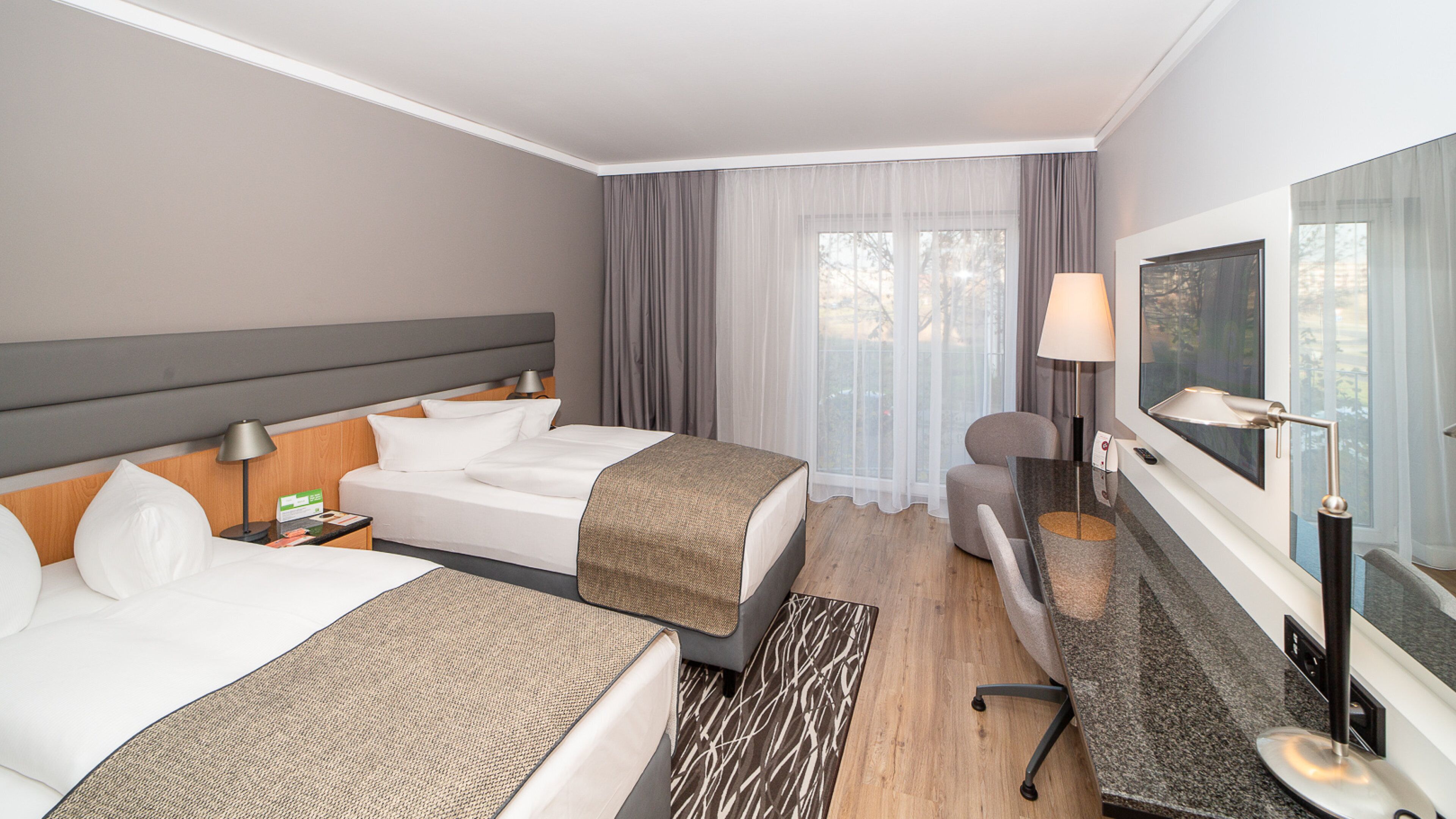 Una habitación de hotel con dos camas, un escritorio, una silla, un televisor y un ventanal con cortinas.