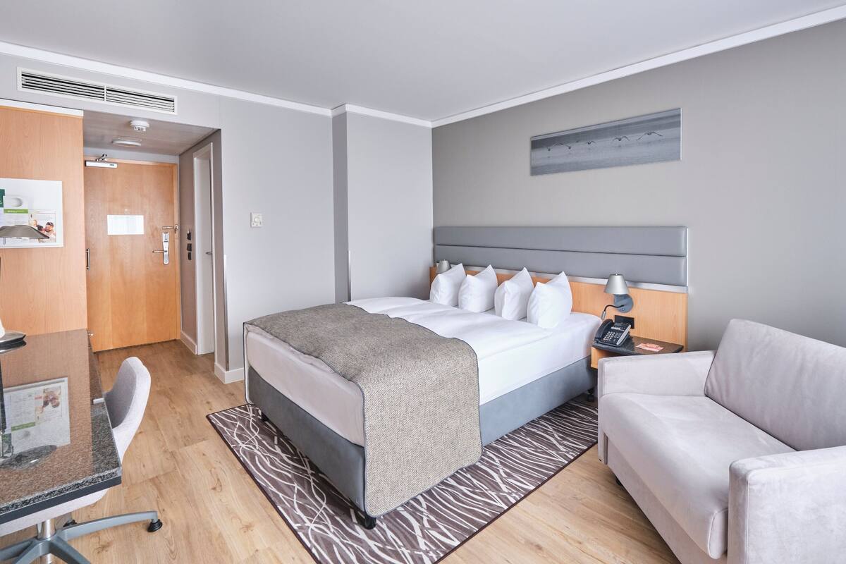 A hotel room with a bed, a desk, a chair, a sofa, and a painting on the wall.