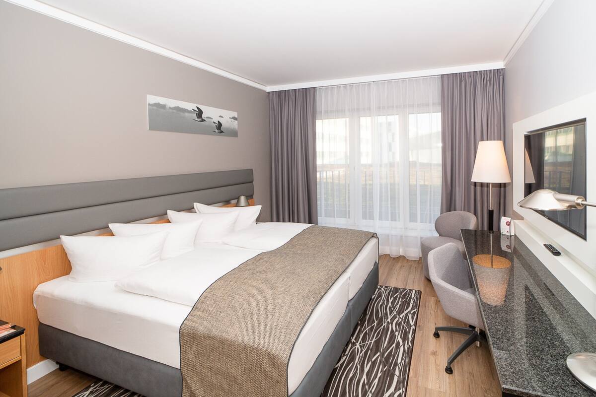 A hotel room with a large bed, a desk, a chair, a TV, and a window with curtains.