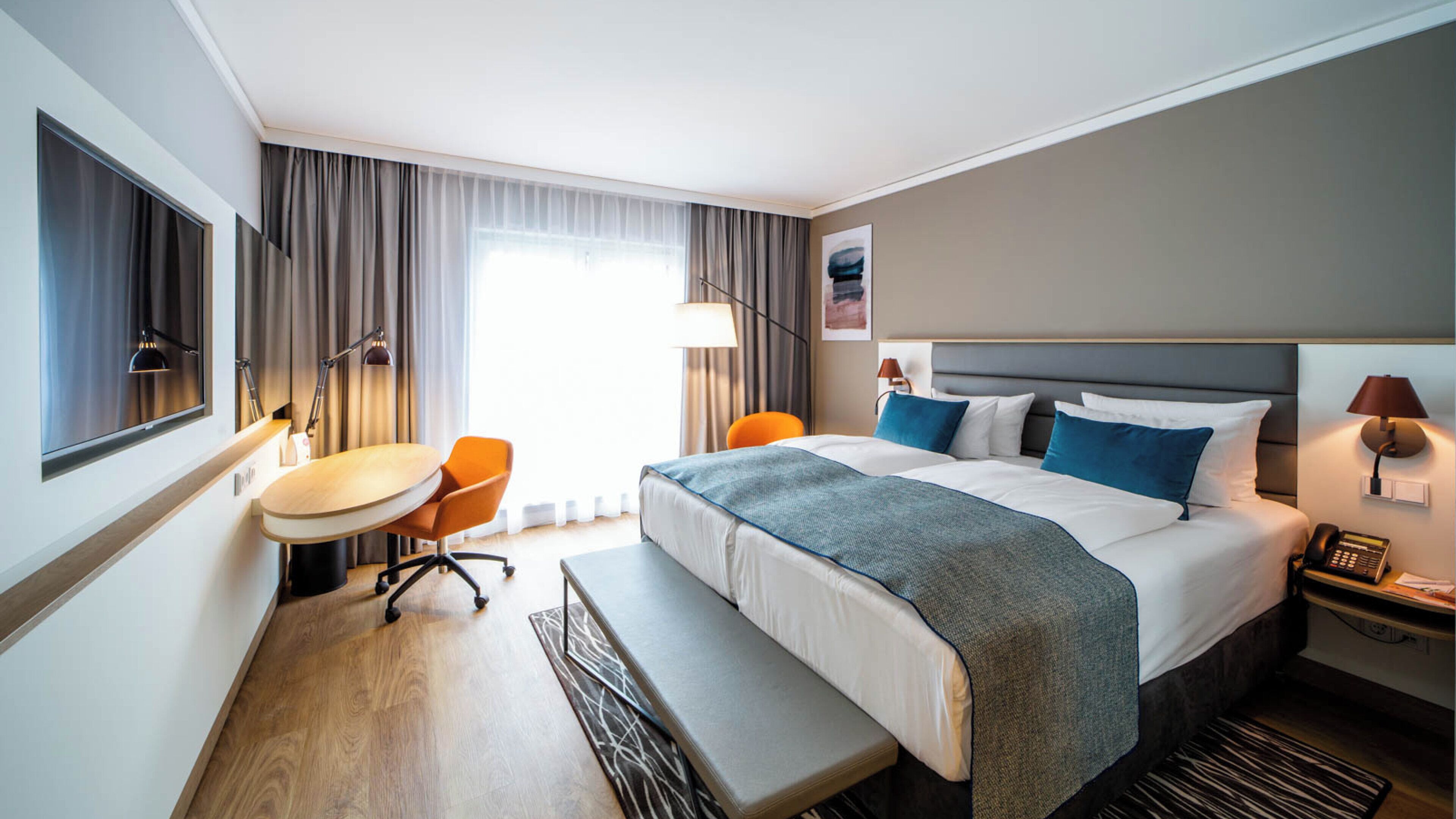 A hotel room with a bed, bedside table, lamp, chair, desk, and a window with curtains.