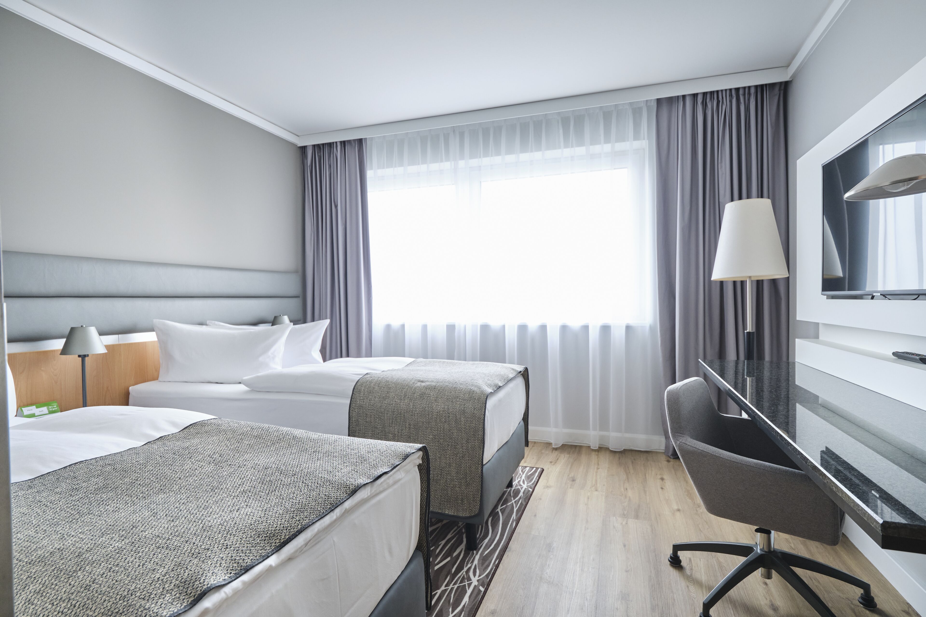 A hotel room with two beds, a desk, a chair, a TV, and a window with curtains.
