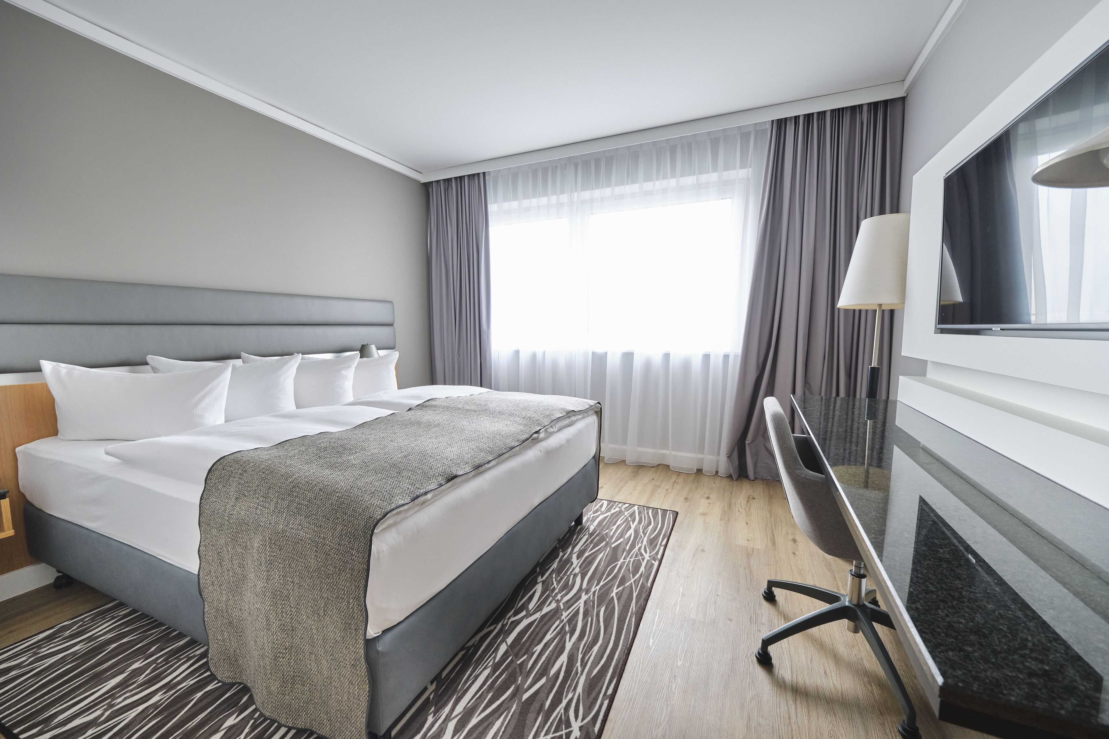 A hotel room with a large bed, a desk, a TV, and a window with curtains.
