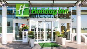 The exterior of a Holiday Inn hotel with a glass entrance, a green 'Holiday Inn' sign, and potted plants at the entrance. - Holiday Inn Berlin Airport - Conference Centre by IHG (Schoenefeld)