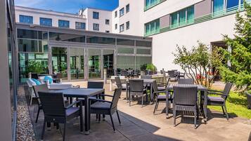 Ein Außenbereich mit Terrasse, Tischen und Stühlen, einer Glaswand und einem Gebäude im Hintergrund.