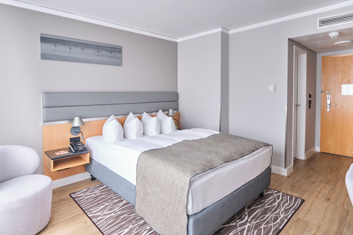 Una camera d'albergo moderna con un letto grande, un comodino con una lampada, una sedia e un pavimento in legno.