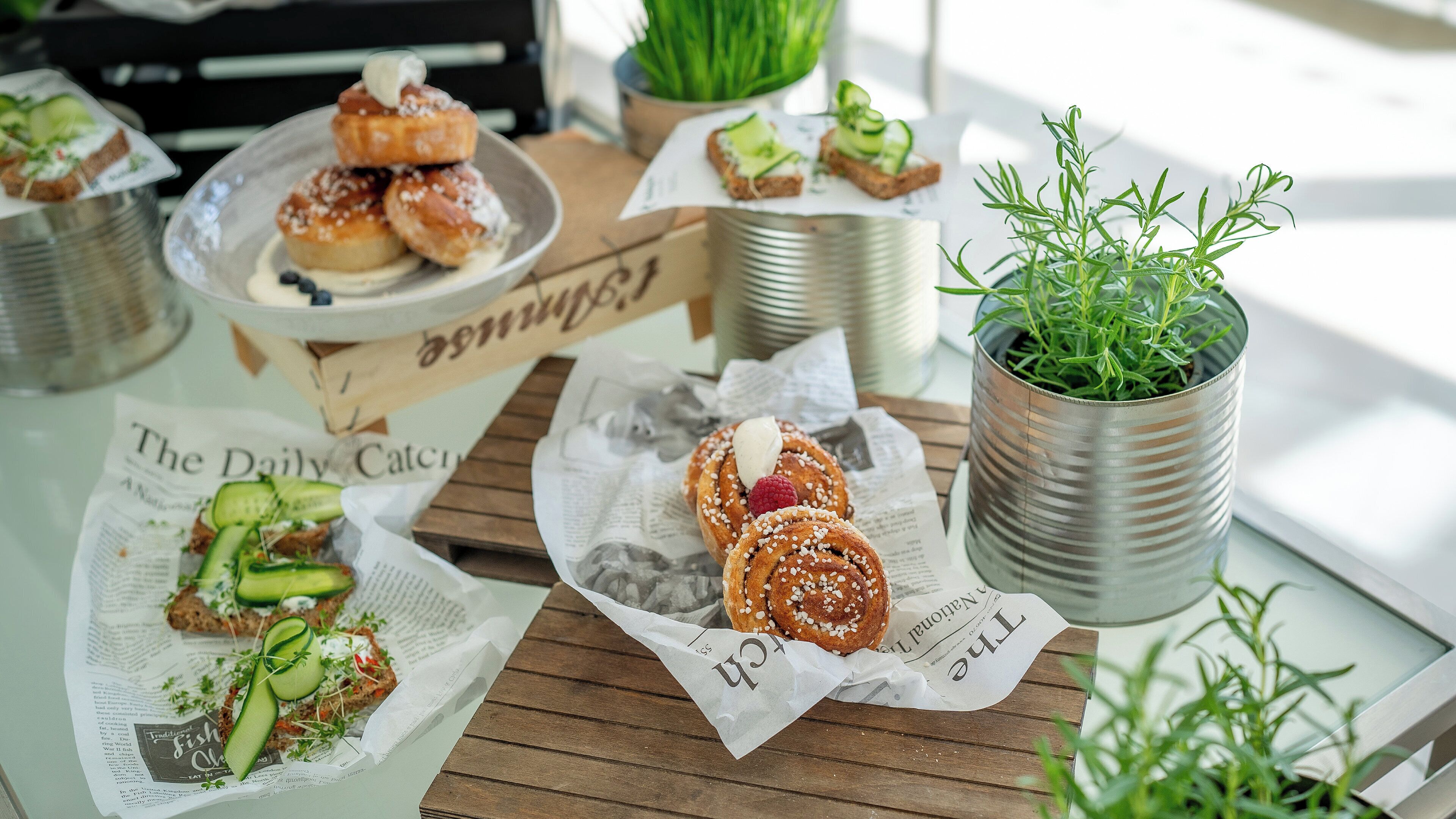 Une table sur laquelle sont disposés divers aliments, notamment des pâtisseries et des sandwichs, le tout présenté sur des feuilles de journal. On peut également voir des récipients en métal et une plante en pot.