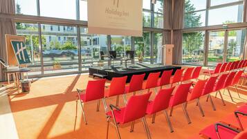 Un salón de conferencias con un escenario, sillas rojas y ventanas amplias que dan a un espacio exterior.