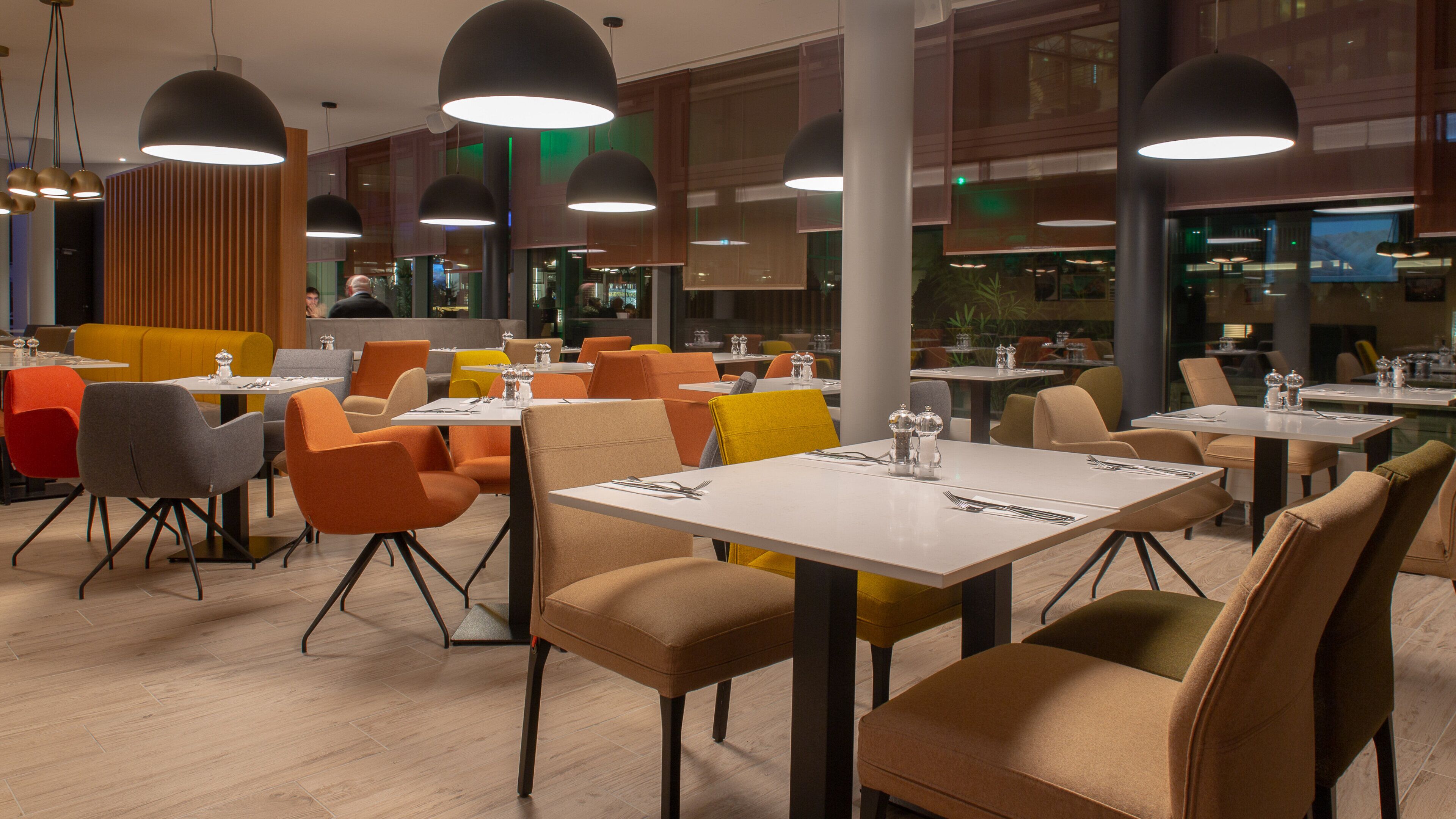 A modern restaurant with multiple tables and chairs, large pendant lights, and a view of the dining area through large windows.