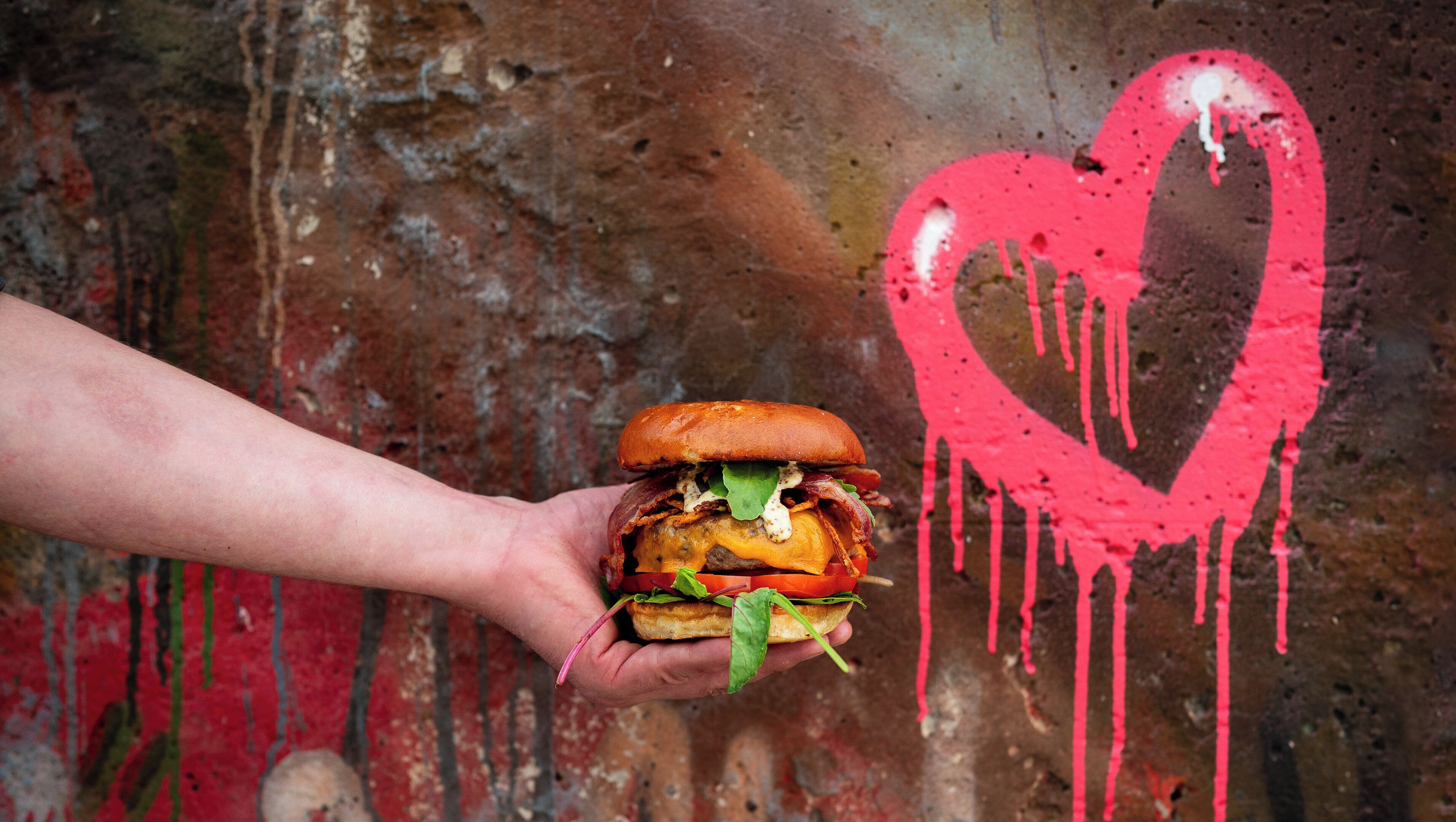 A hand holding a burger with a heart-shaped graffiti in the background.