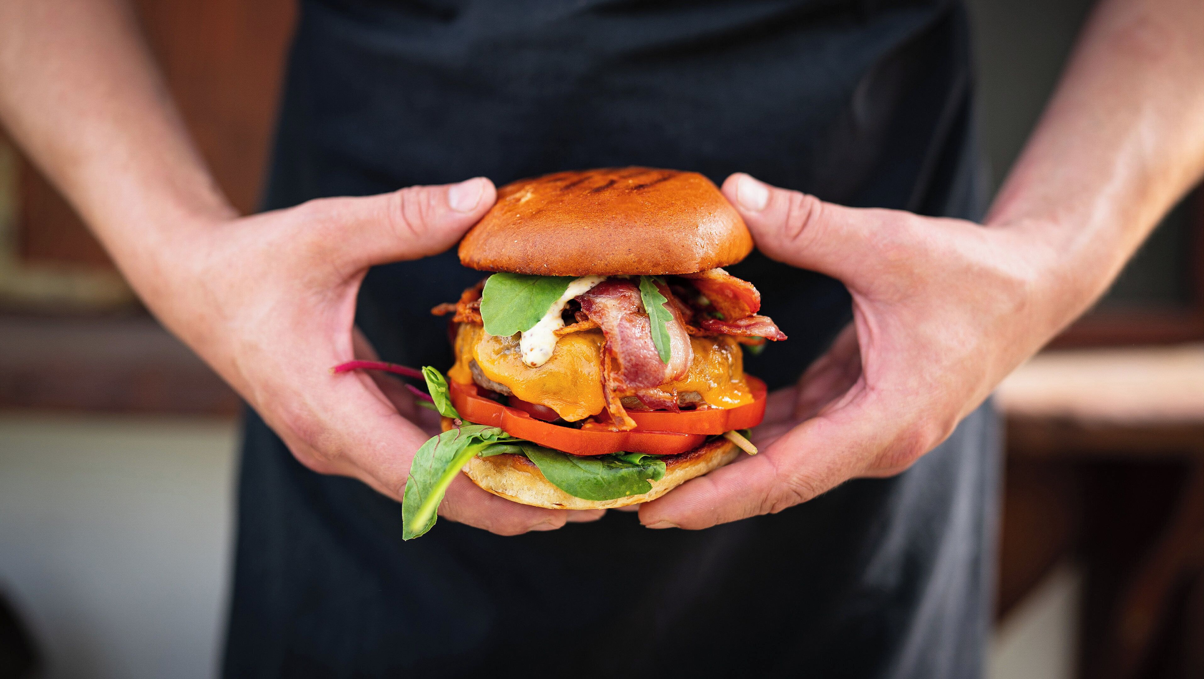 A person holding a burger with visible ingredients like lettuce, tomato, cheese, and bacon.