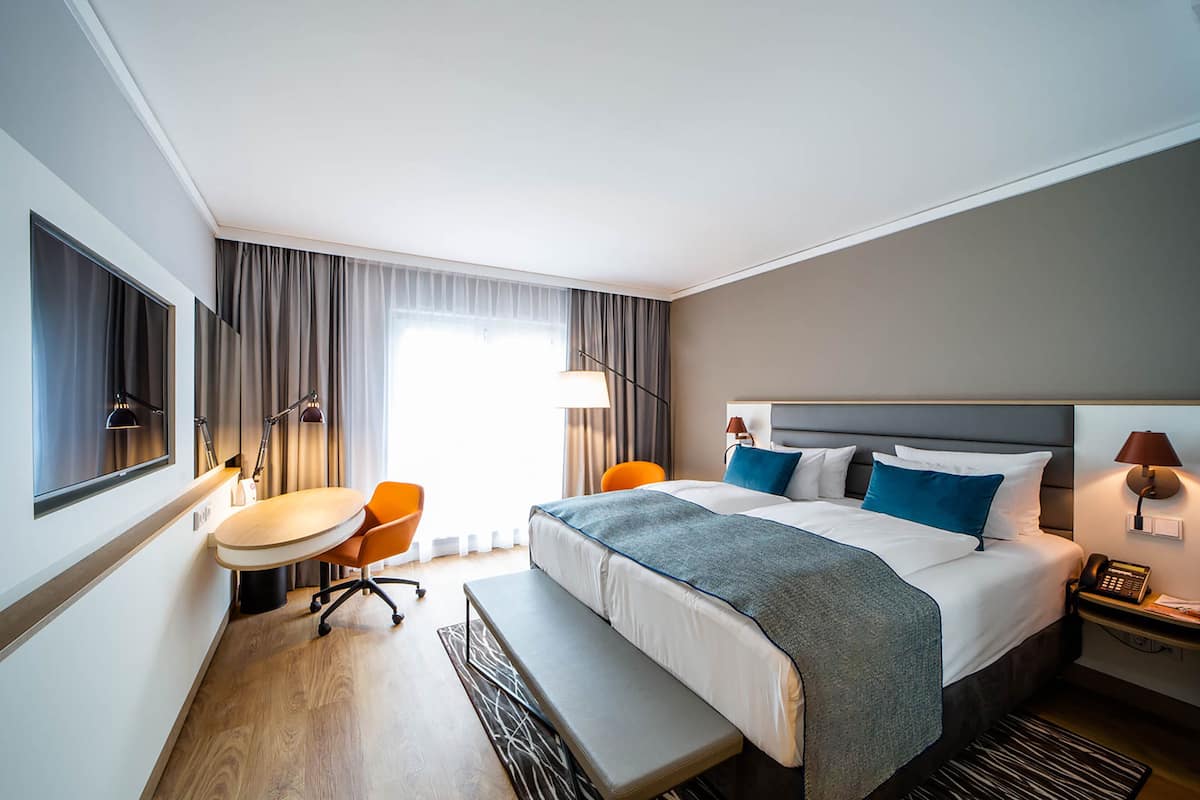 A hotel room with a large bed, a desk, two chairs, a bench, a lamp, and a telephone.