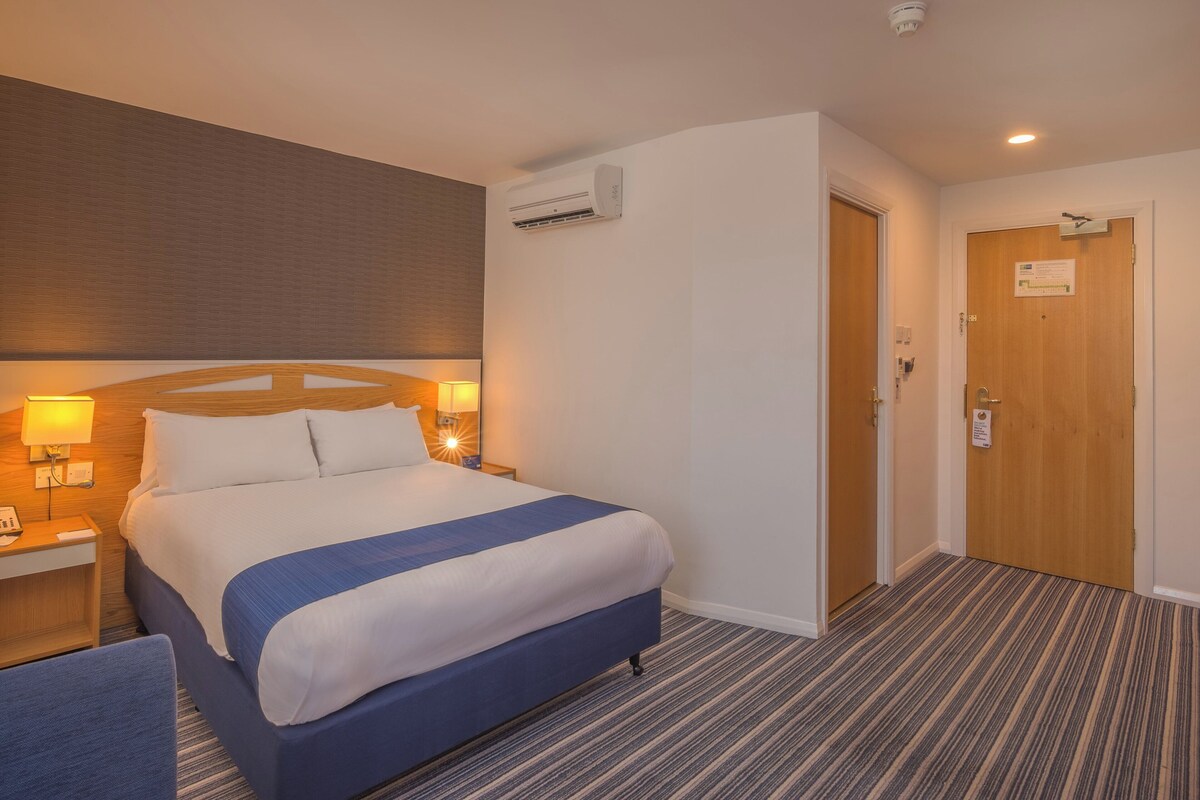 A hotel room with a bed, bedside table, lamp, and a door with a sign.