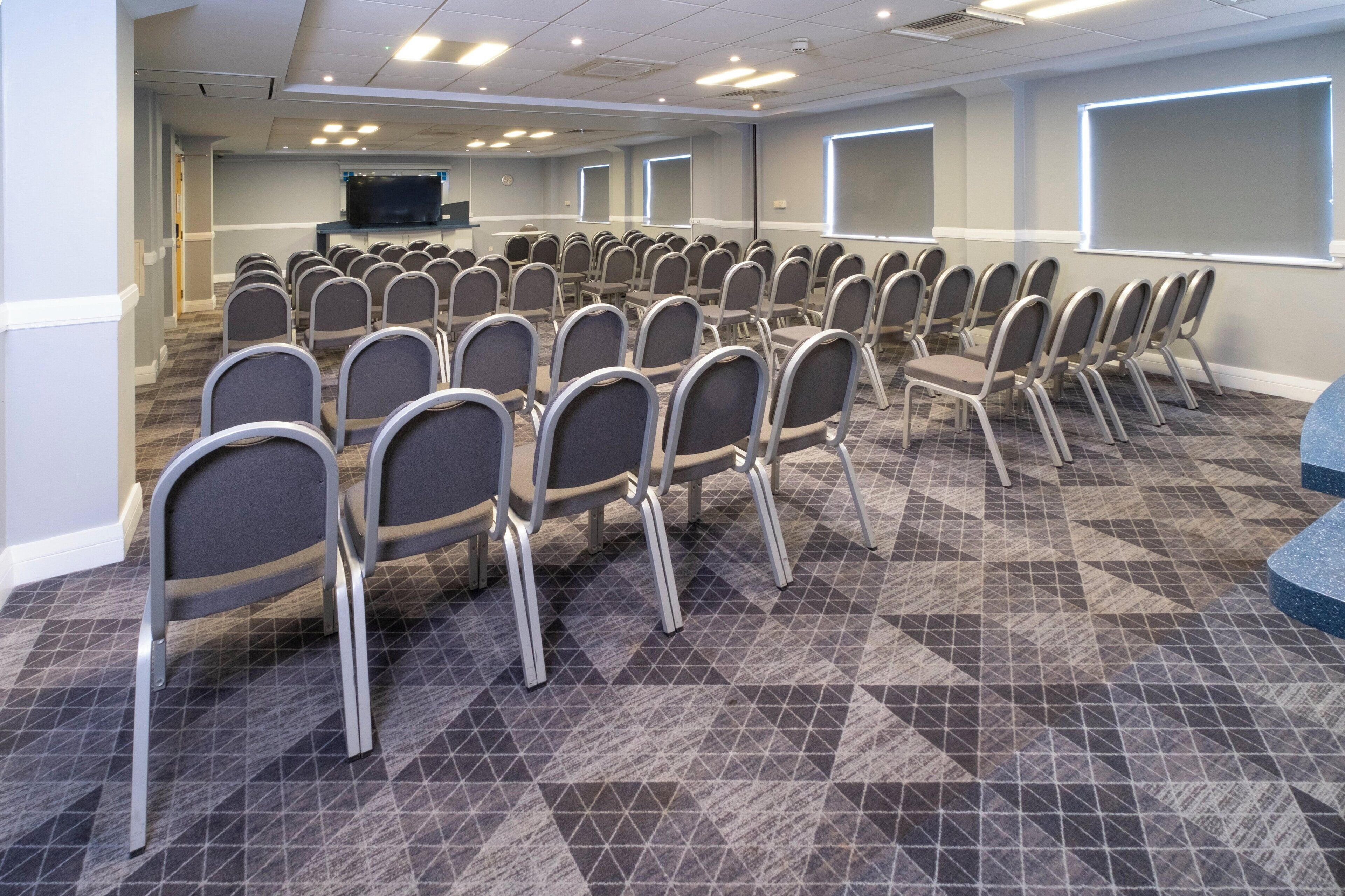 Un salón de conferencias con hileras de sillas plegables, una pantalla grande y suelo alfombrado.