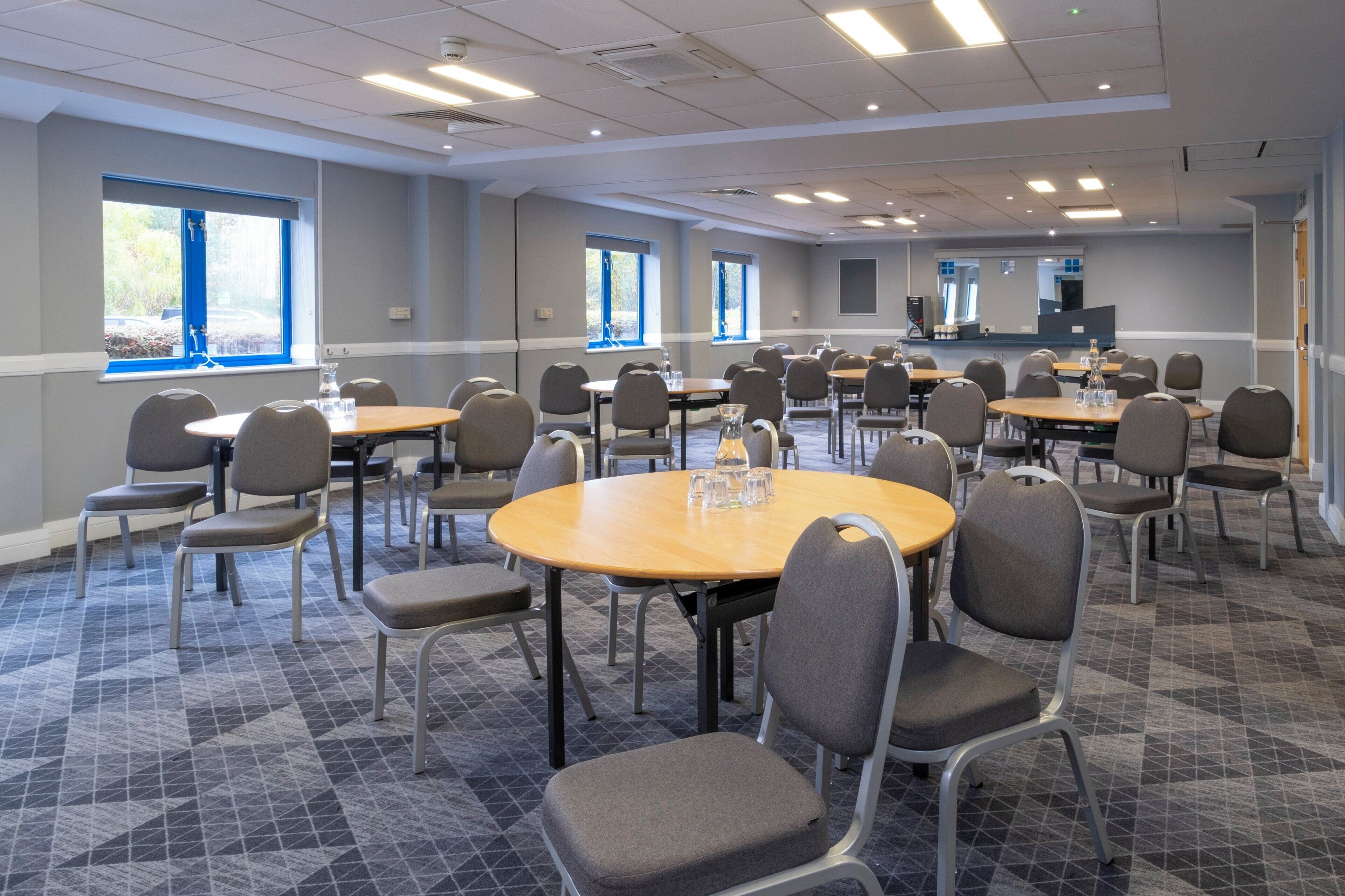 A conference room with multiple tables and chairs, large windows, and a carpeted floor.