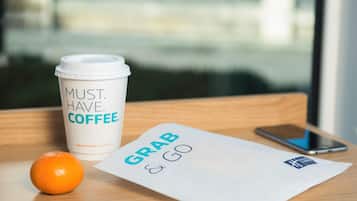 Eine Kaffeetasse mit Schriftzug, ein Papier mit der Aufschrift 'GRAB & GO', ein Smartphone und eine Orange auf einer Holztischplatte.