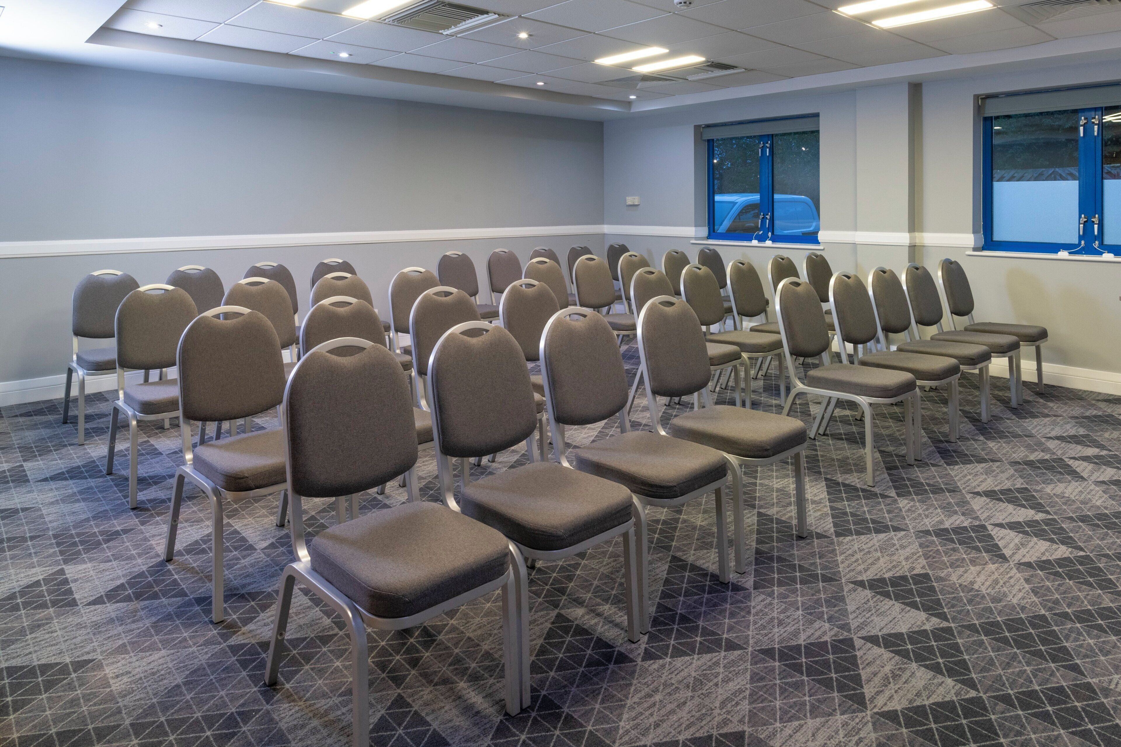 Un salón de conferencias con filas de sillas, una alfombra estampada y ventanas con marcos azules.