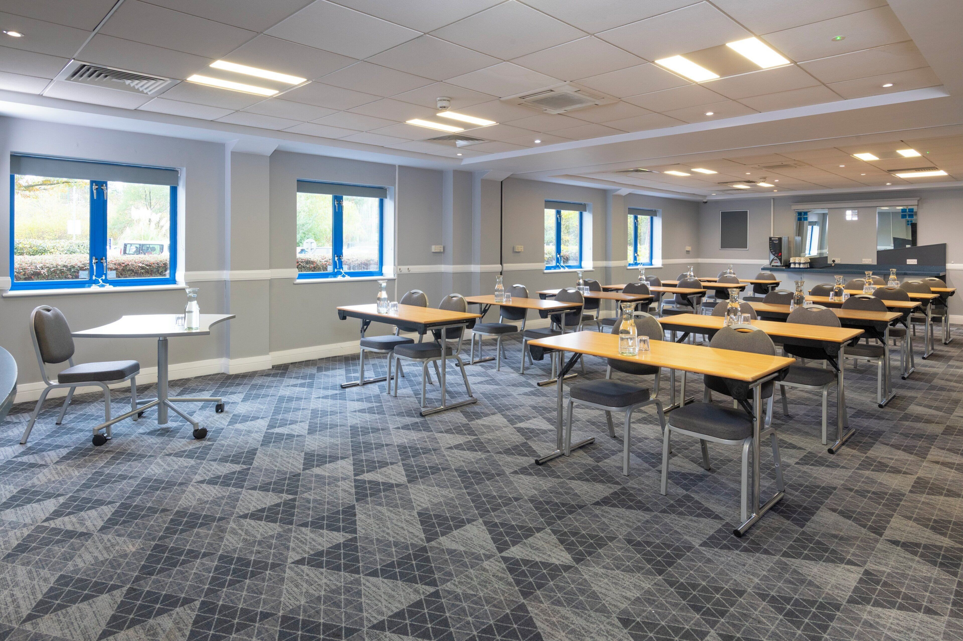 A classroom with rows of desks and chairs, large windows, and a projector screen.