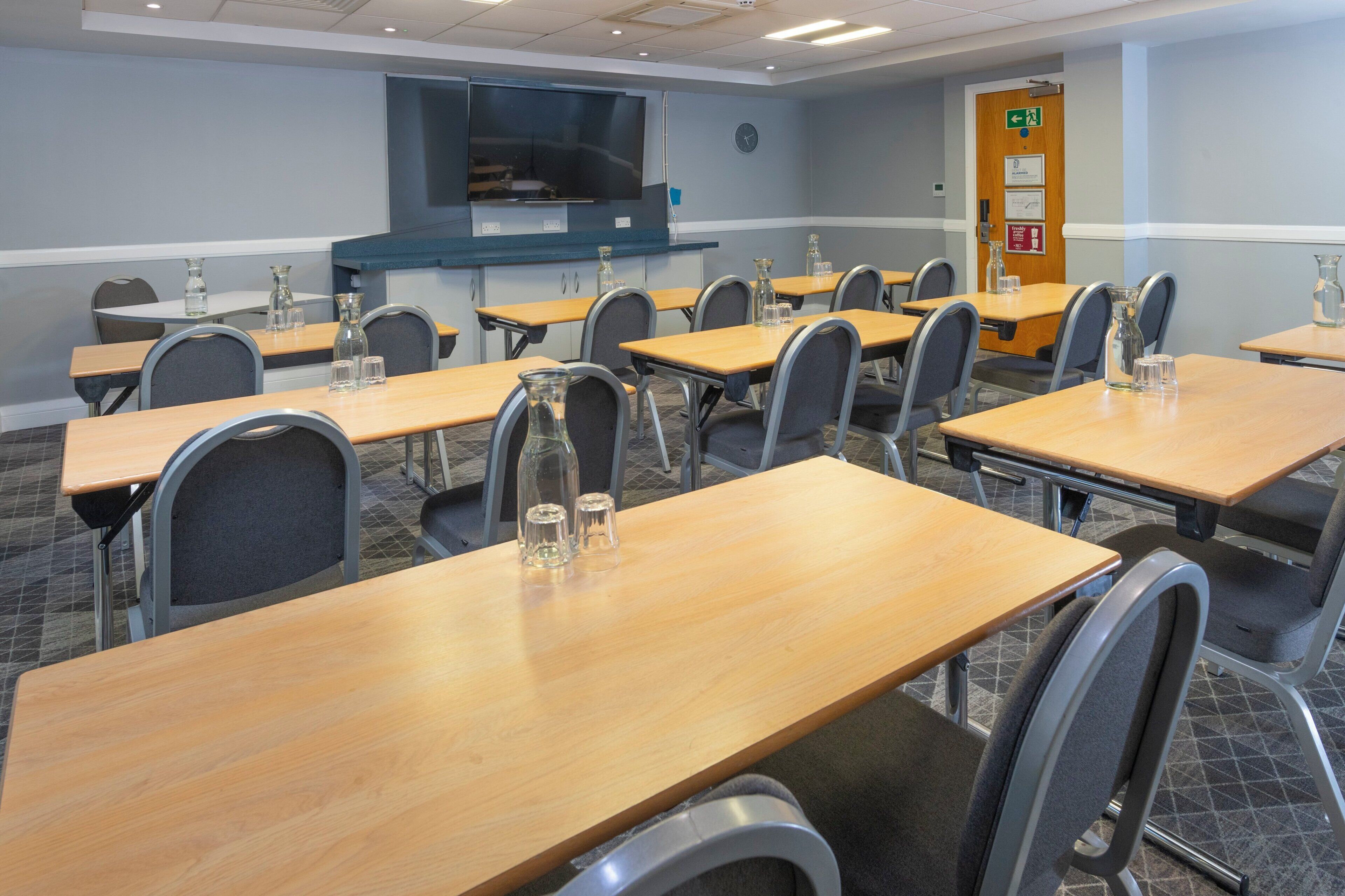 A conference room with multiple tables and chairs, a large screen, and a door with emergency signage.