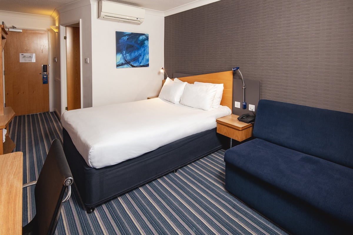 Une chambre d’hôtel avec un lit, une table de chevet avec une lampe, un canapé bleu et un tapis à rayures.