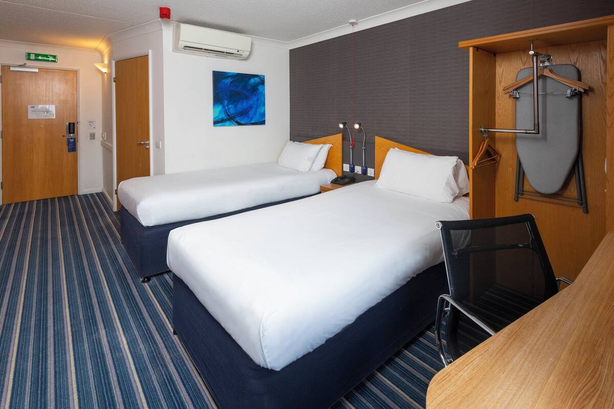 Une chambre d’hôtel avec deux lits, un bureau, une chaise, une planche à repasser et un tableau au mur.