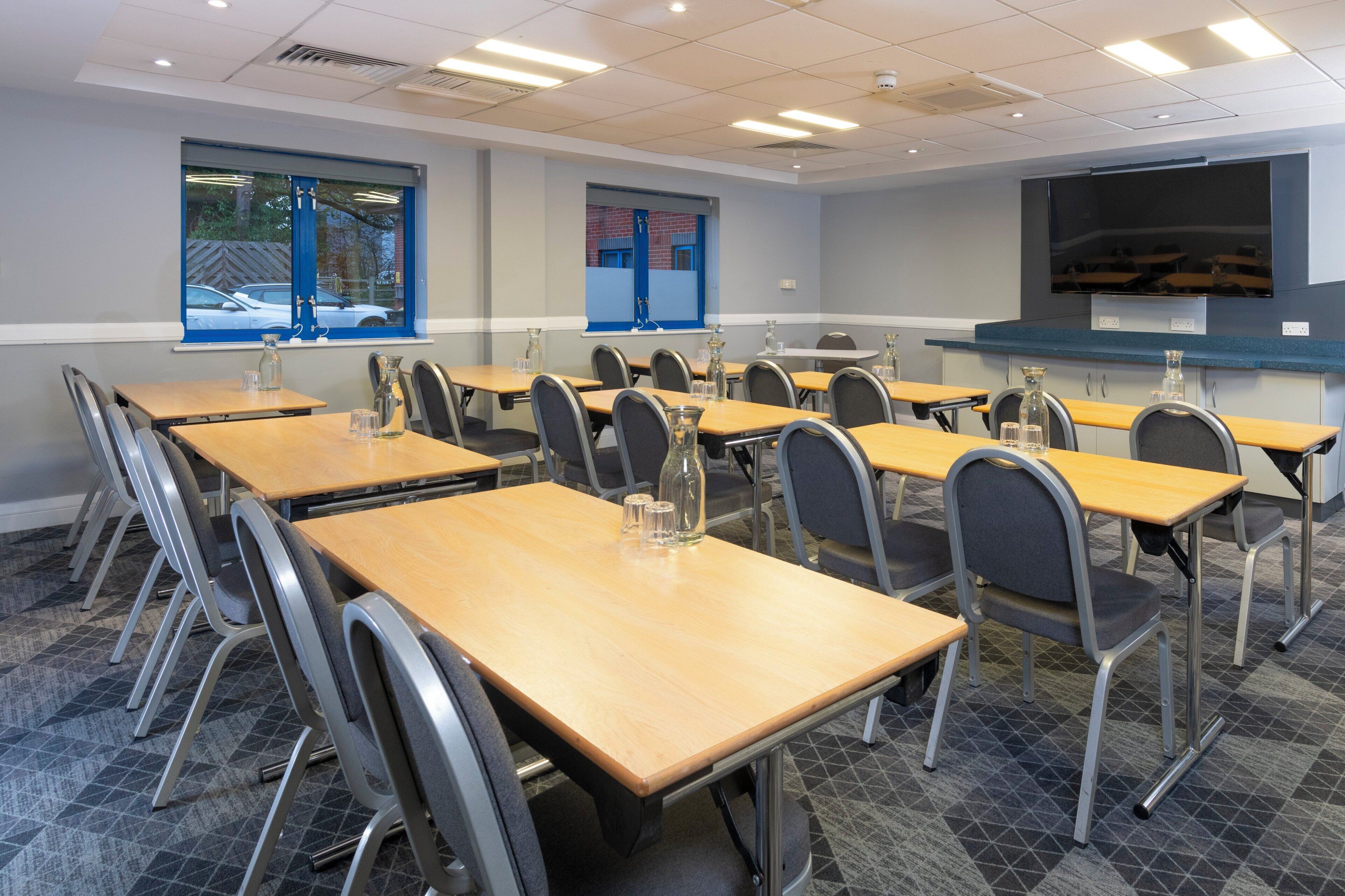 A conference room with multiple tables and chairs, a large screen, and windows with blue frames.