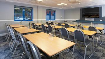 A conference room with multiple tables and chairs, a large screen, and windows with blue frames.