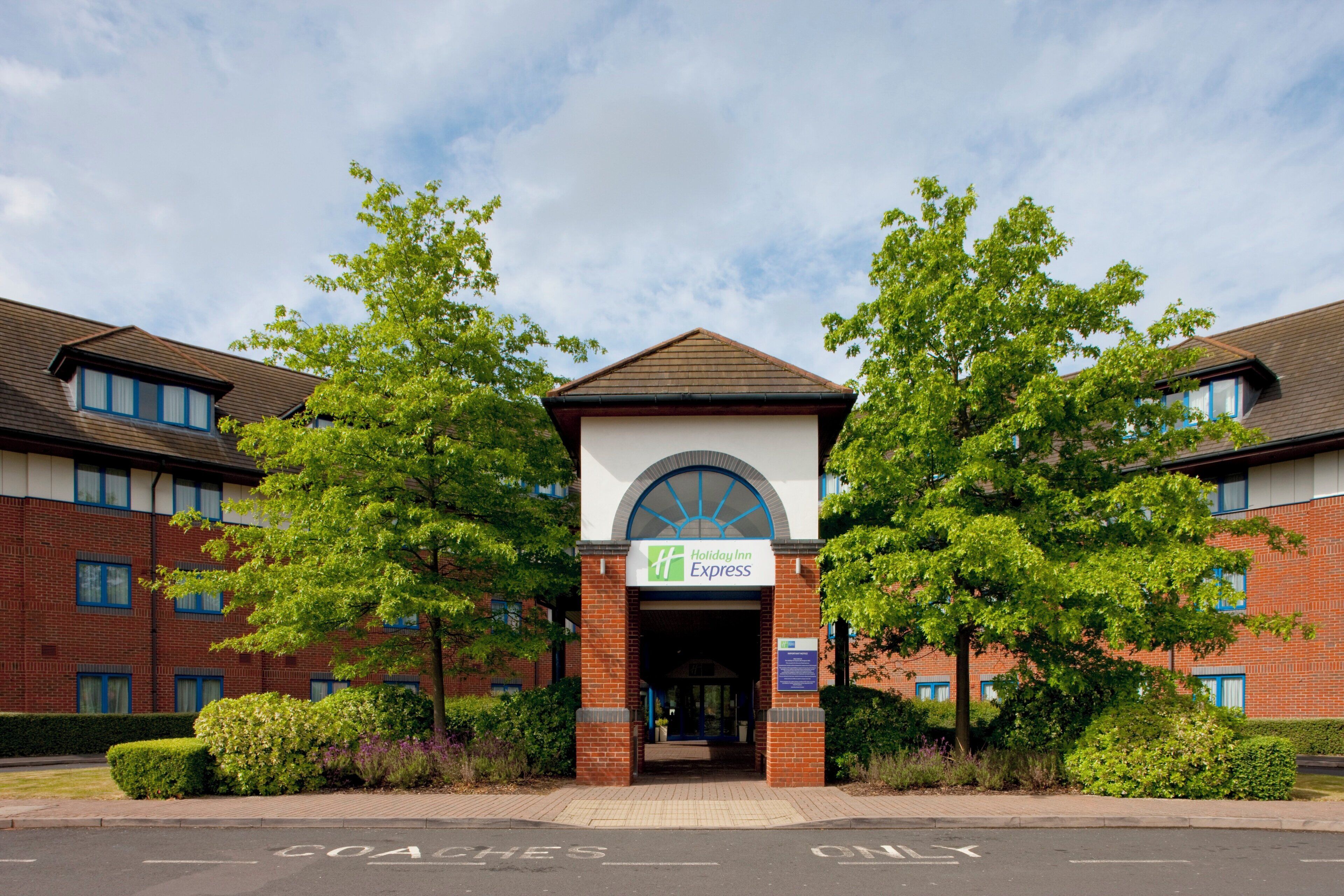 A Holiday Inn Express building with a brick facade, a central entrance with a large arched window, and surrounding greenery.
