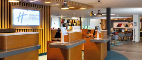 A Holiday Inn Express reception area with wooden reception desks, a large illuminated sign, and a seating area with tables and chairs.