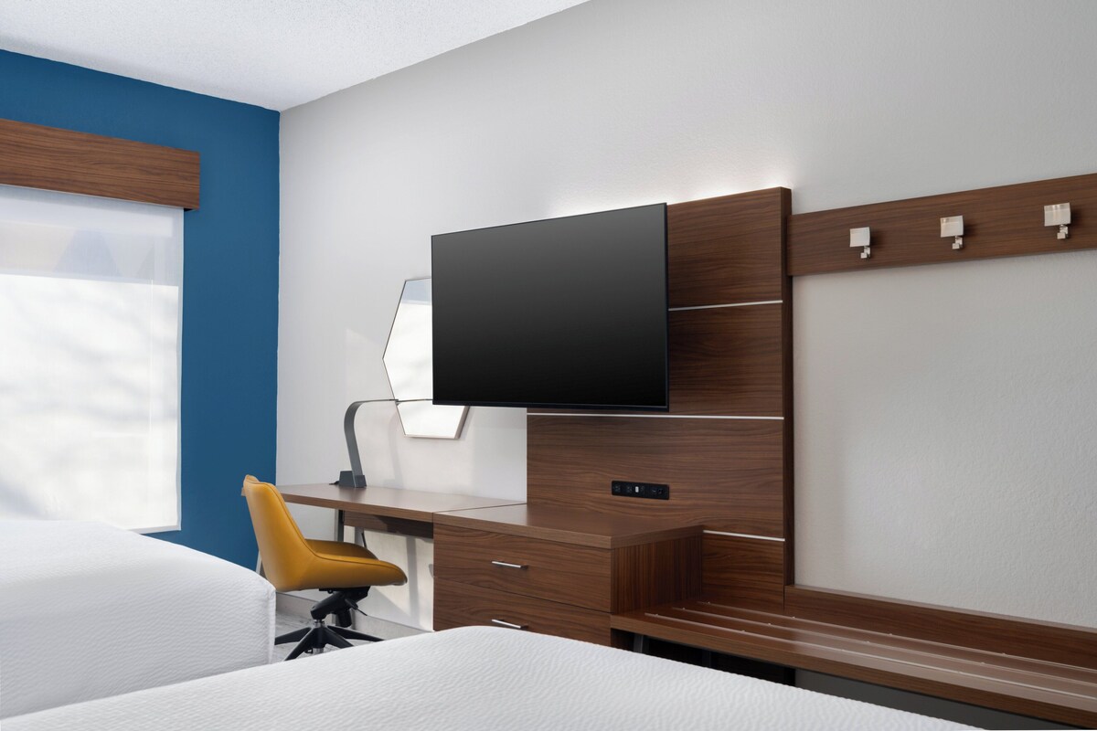 Habitación de hotel con cama, escritorio con silla, televisor de pantalla plana en la pared y ventana con persianas.