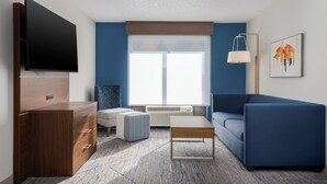 Un salon moderne avec un téléviseur à écran plat, un meuble en bois, un canapé bleu, un petit pouf, une table basse et une fenêtre avec des stores.