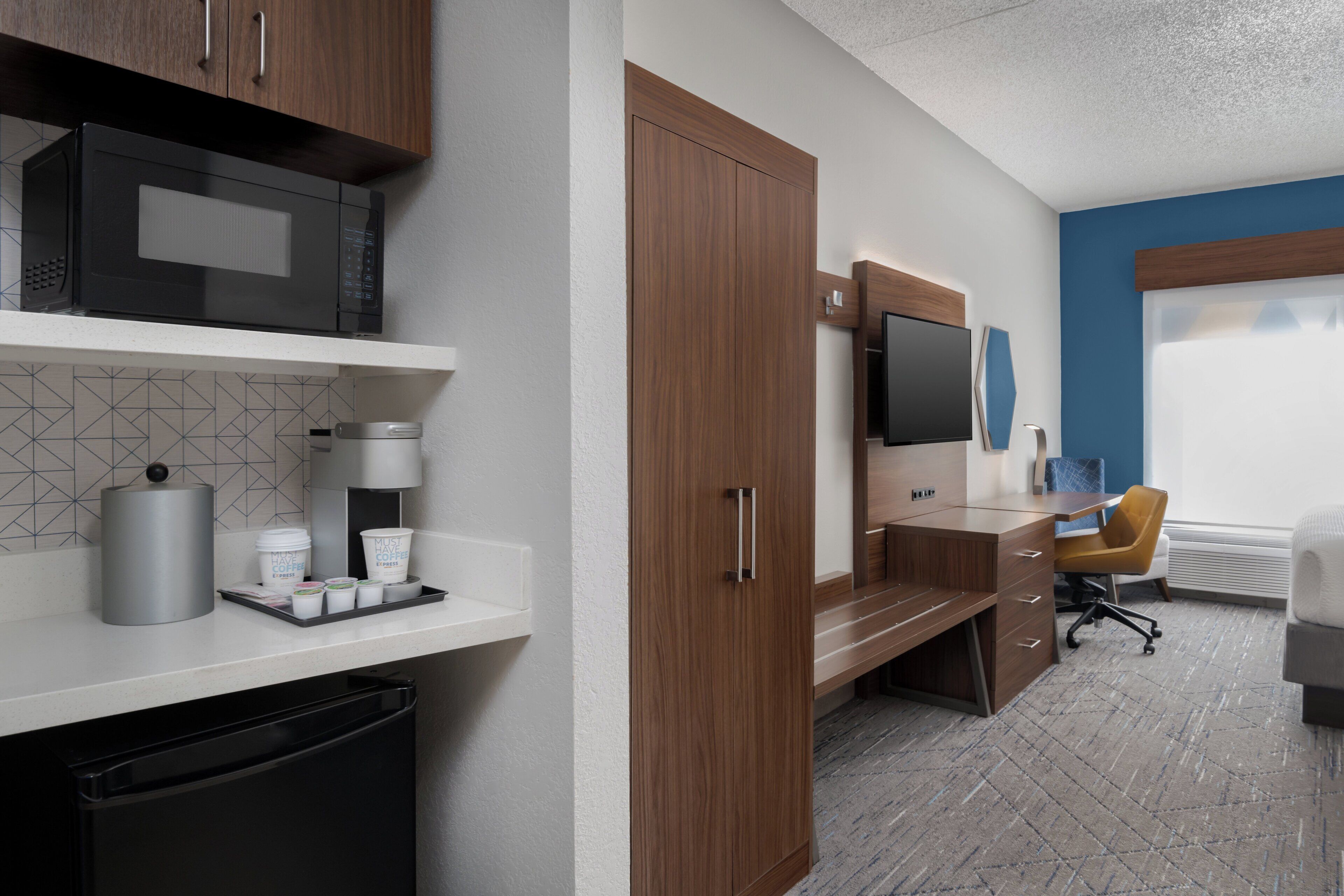 Una habitación de hotel moderna con kitchenette, un escritorio, una silla y un ventanal grande.