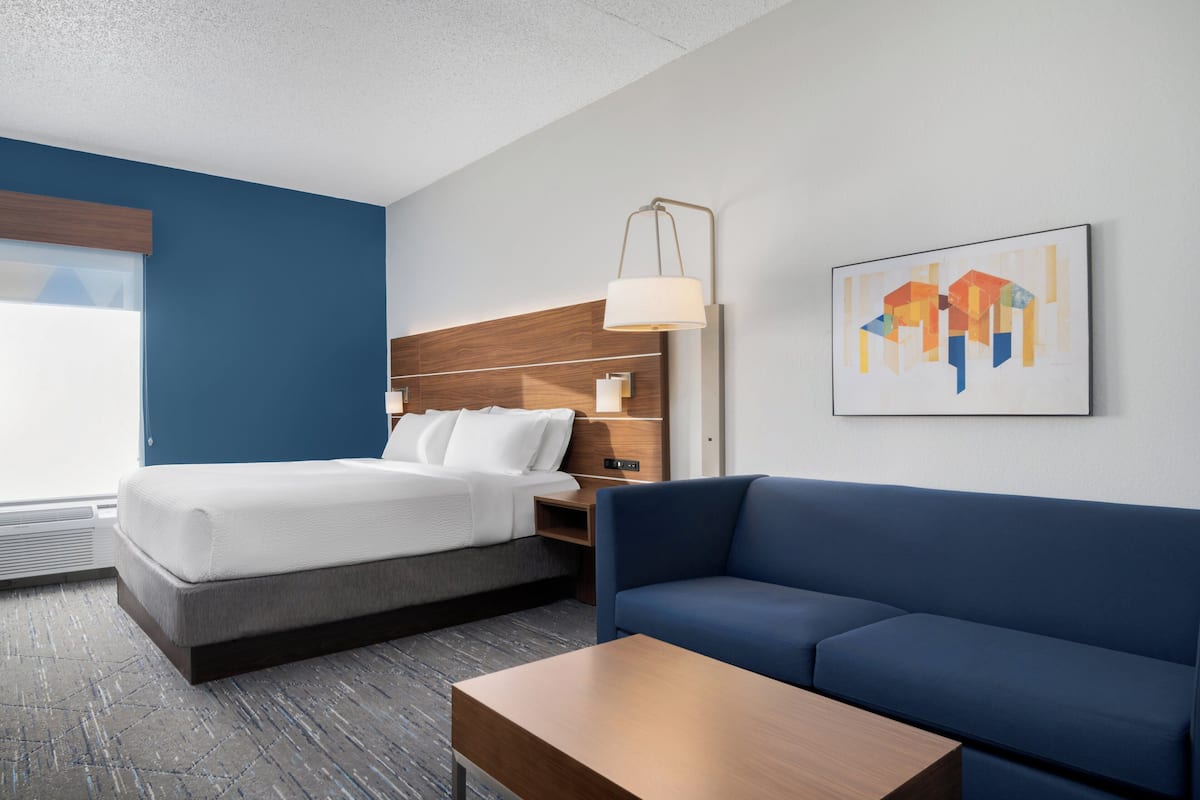 Una habitación de hotel con una cama, un sofá azul, una mesita de noche de madera, una lámpara y un cuadro en la pared.