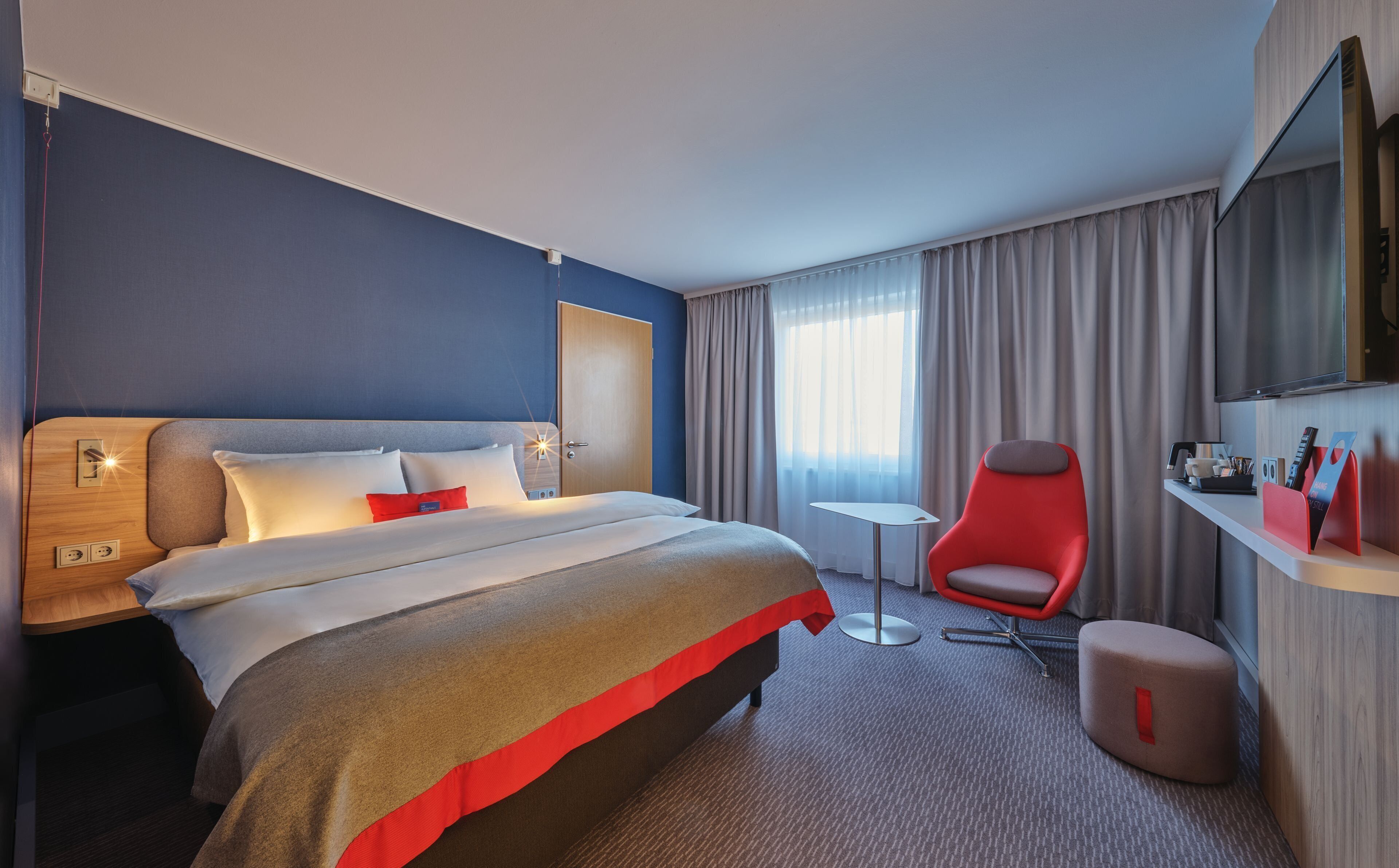 Quarto de hotel com cama, uma cadeira vermelha, uma mesinha, televisão e janela com cortinas.