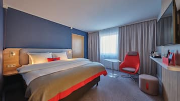 Quarto de hotel com cama, uma cadeira vermelha, uma mesinha, televisão e janela com cortinas.