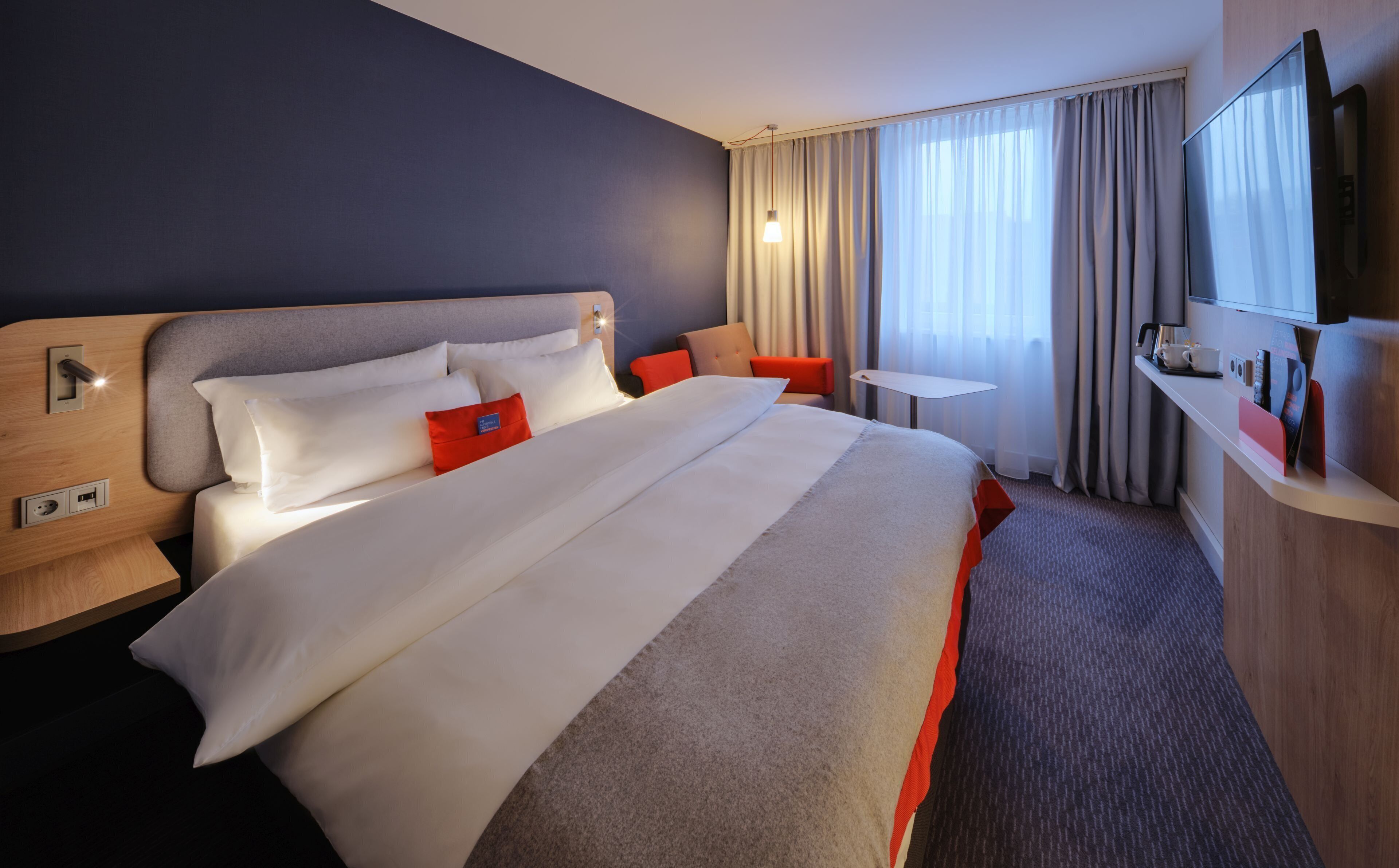 Quarto de hotel com uma cama grande, uma escrivaninha, uma cadeira, uma televisão e uma janela com cortinas.