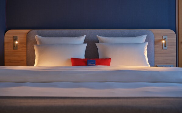 A neatly made bed with white linens and two pillows, a red accent pillow, and a blue tag in the center. - Holiday Inn Express Köln-Troisdorf by IHG (Troisdorf)