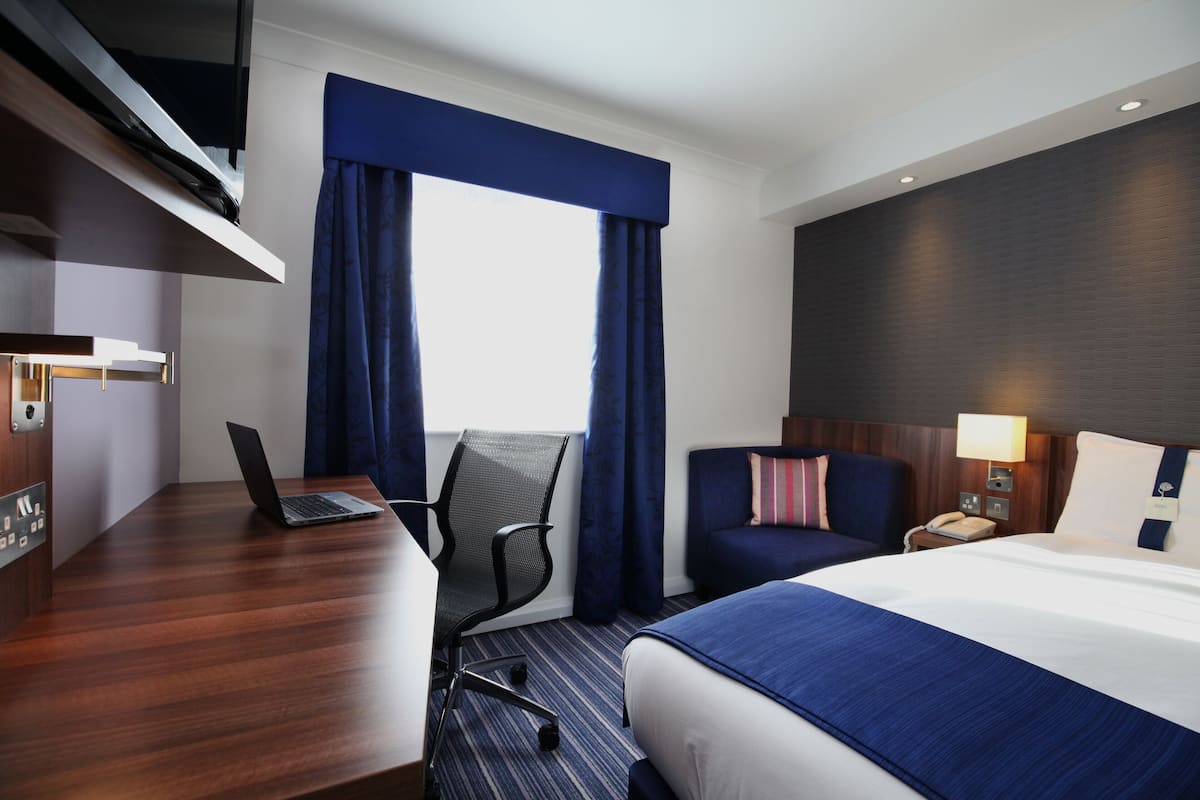 A hotel room with a bed, a desk with a laptop, a chair, a nightstand with a lamp, and a window with curtains.