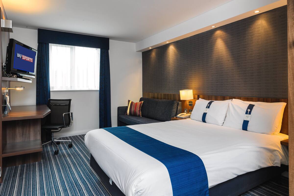 A hotel room with a large bed, a desk with a TV, a chair, and a window with curtains.