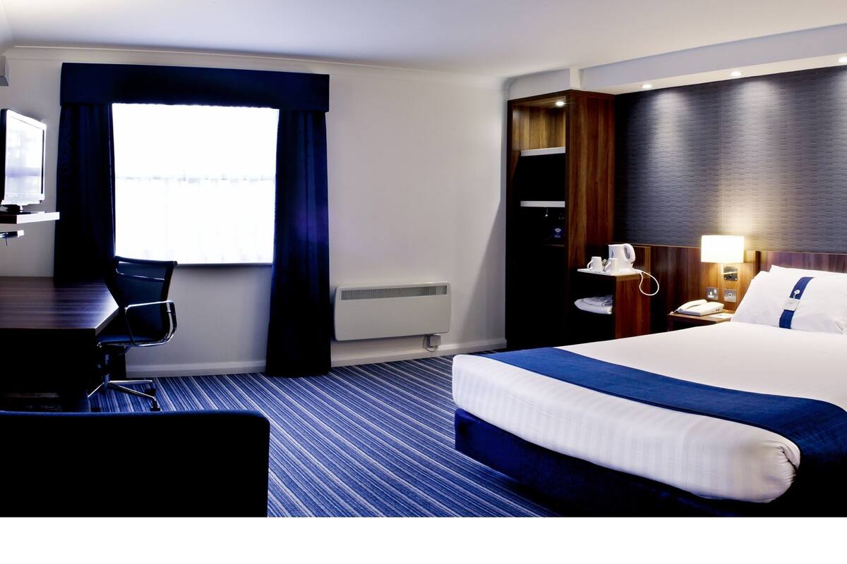 A hotel room with a large bed, a desk with a chair, a nightstand with a lamp, and a view of the outdoors through the window.