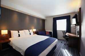 A hotel room with a large bed, a desk, a chair, a TV, and a window with curtains.