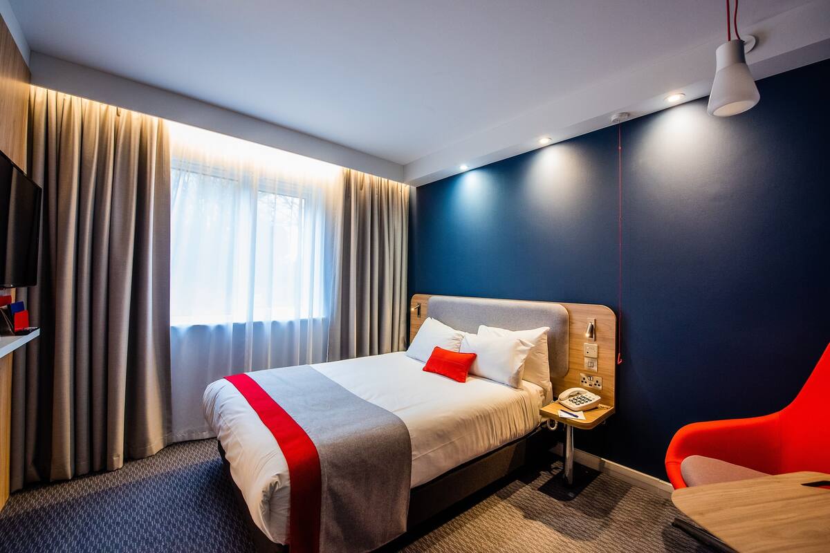 A hotel room with a bed, a nightstand, a desk, a chair, and a window with curtains.