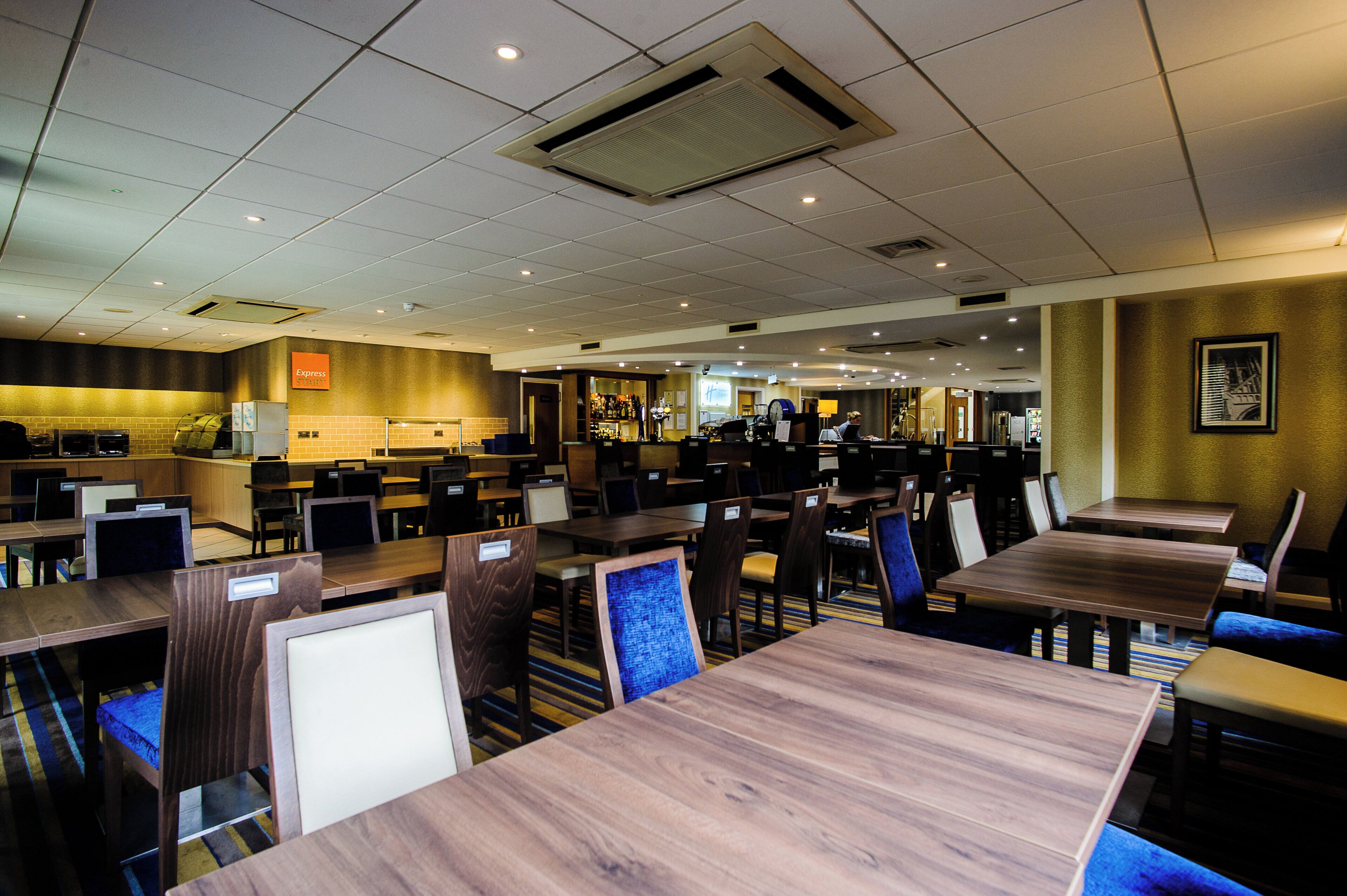 A hotel dining area with multiple tables and chairs, a bar area, and a kitchen in the background.