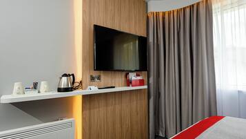 A hotel room with a flat-screen TV mounted on a wooden wall, a white shelf with a kettle, a remote control, and a magazine rack.